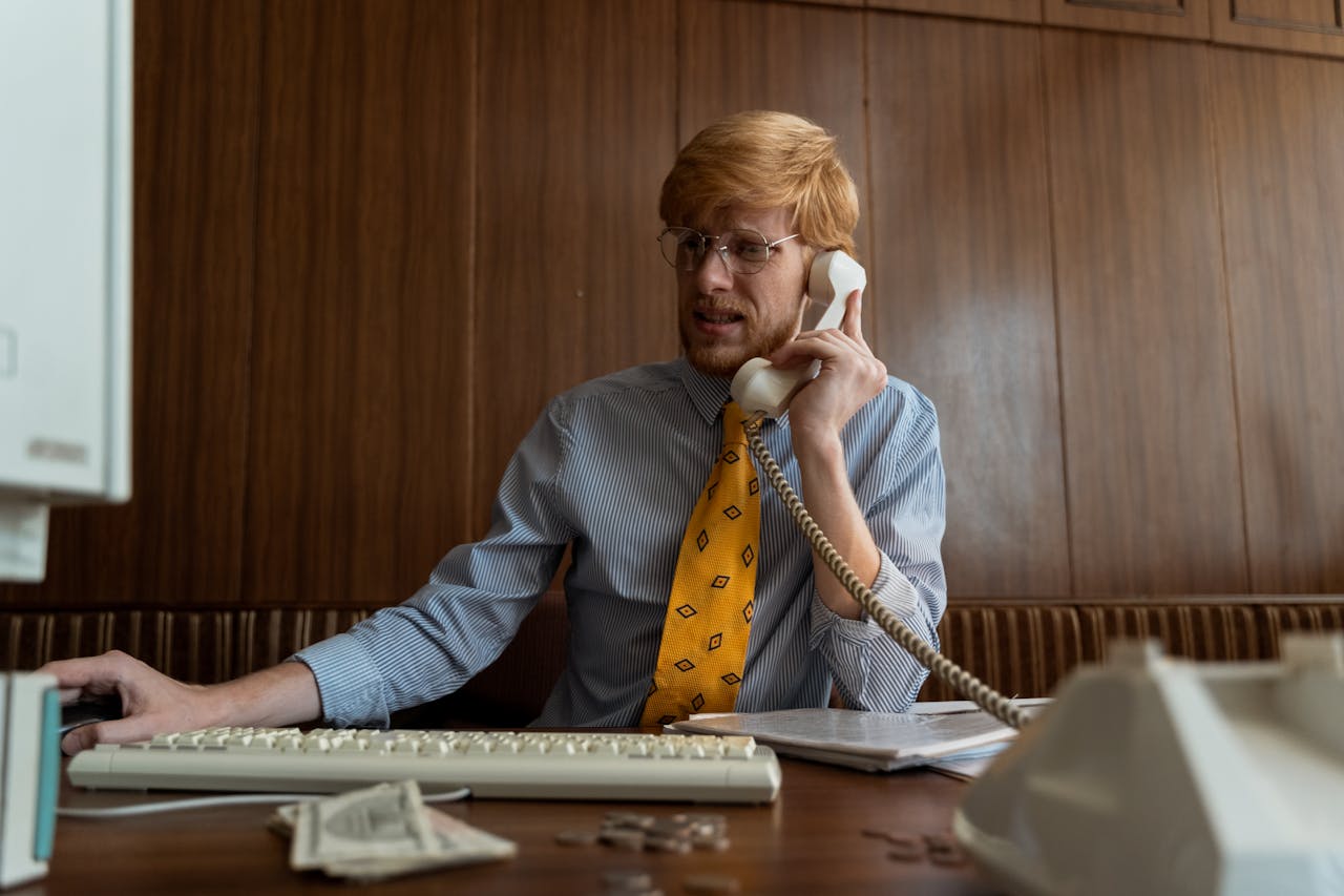 mobile-01 A man in business attire using a vintage telephone and computer in an office setting.